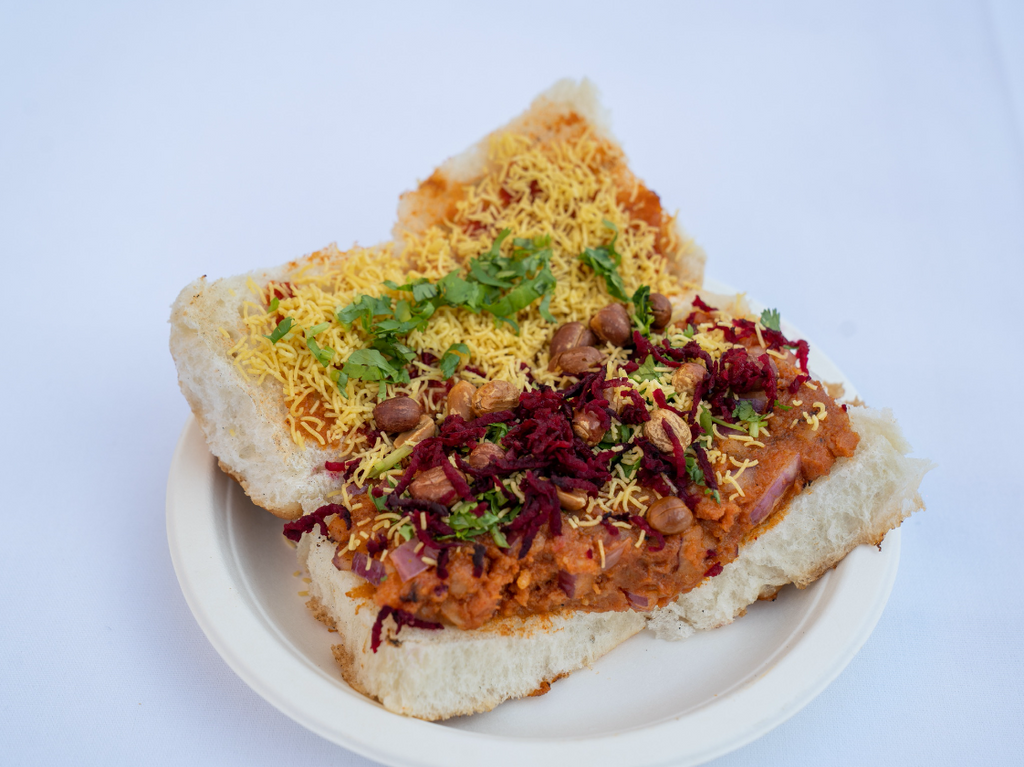 Spicy and tangy potato filling stuffed in a bun with peanuts, onions, dabeli chutney, and thin chickpea flour noodles (Sev) 